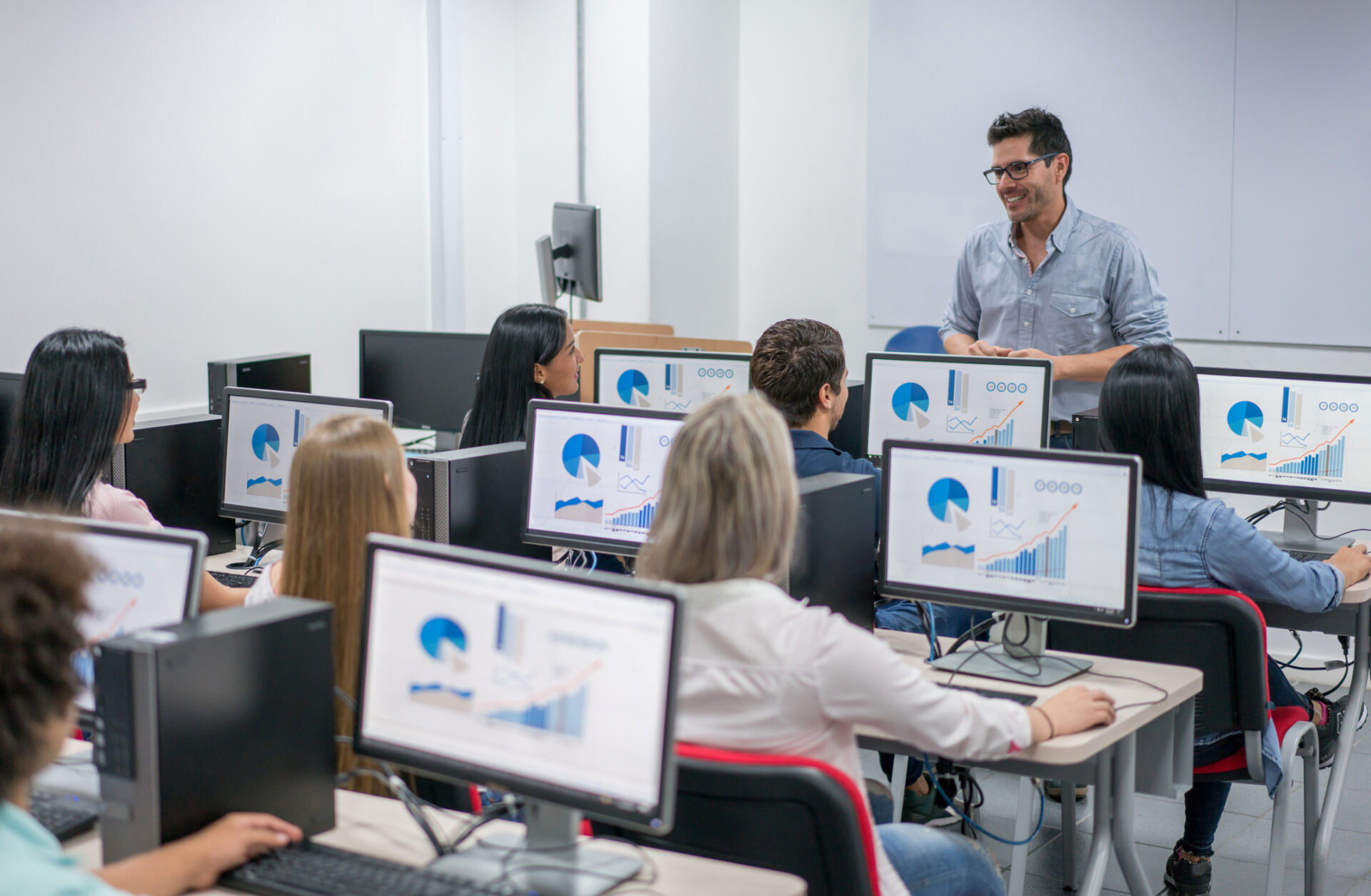 Teacher giving an IT class at school to a group of students Happy teacher giving an IT class at school to a group of students using computers - education concepts. Image on screens were made from scratch by us.